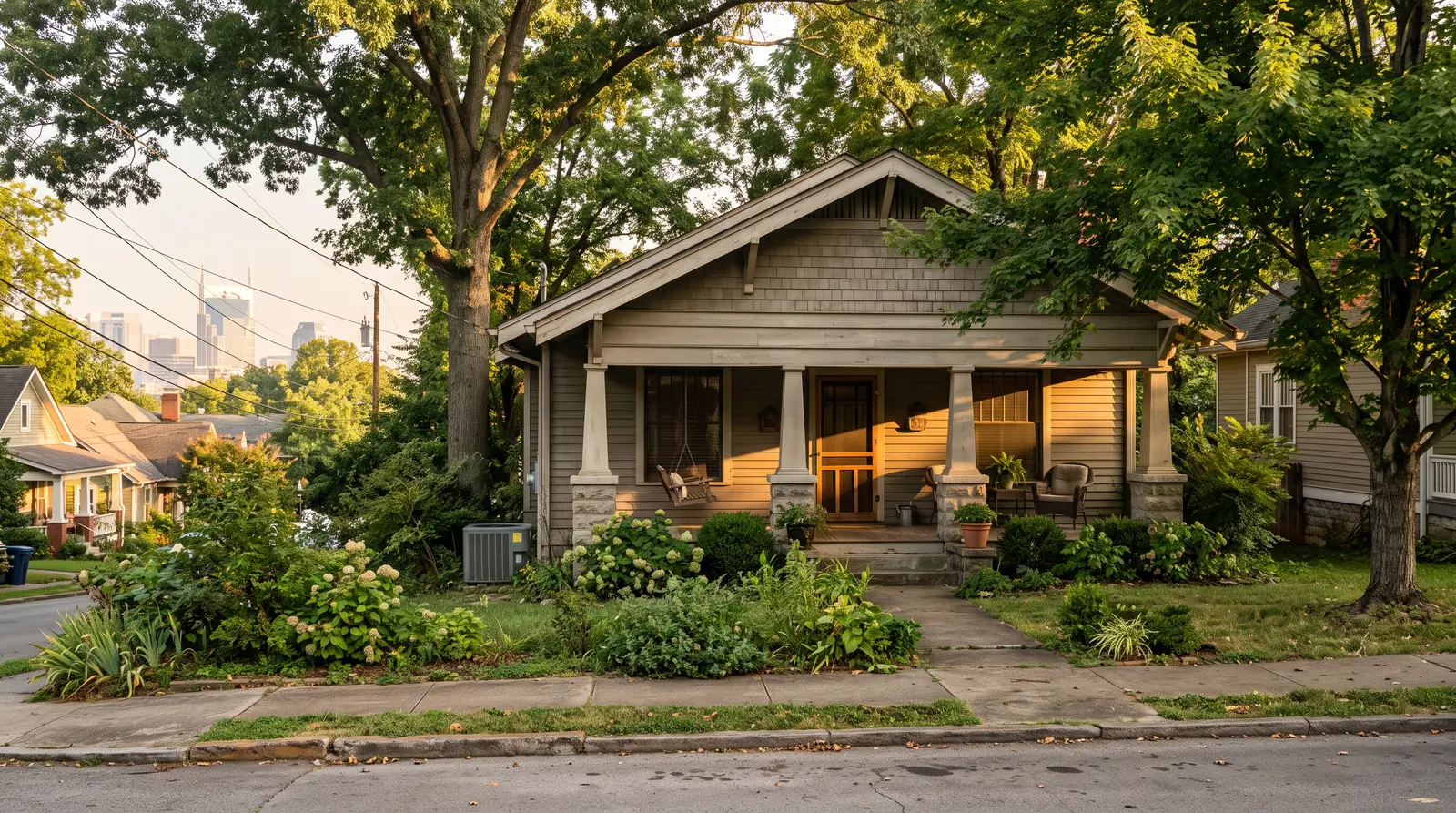 A charming East Nashville craftsman bungalow at golden hour with a deep porch, mature trees and a discreet outdoor condenser tucked beside the foundation, downtown buildings faintly visible far behind