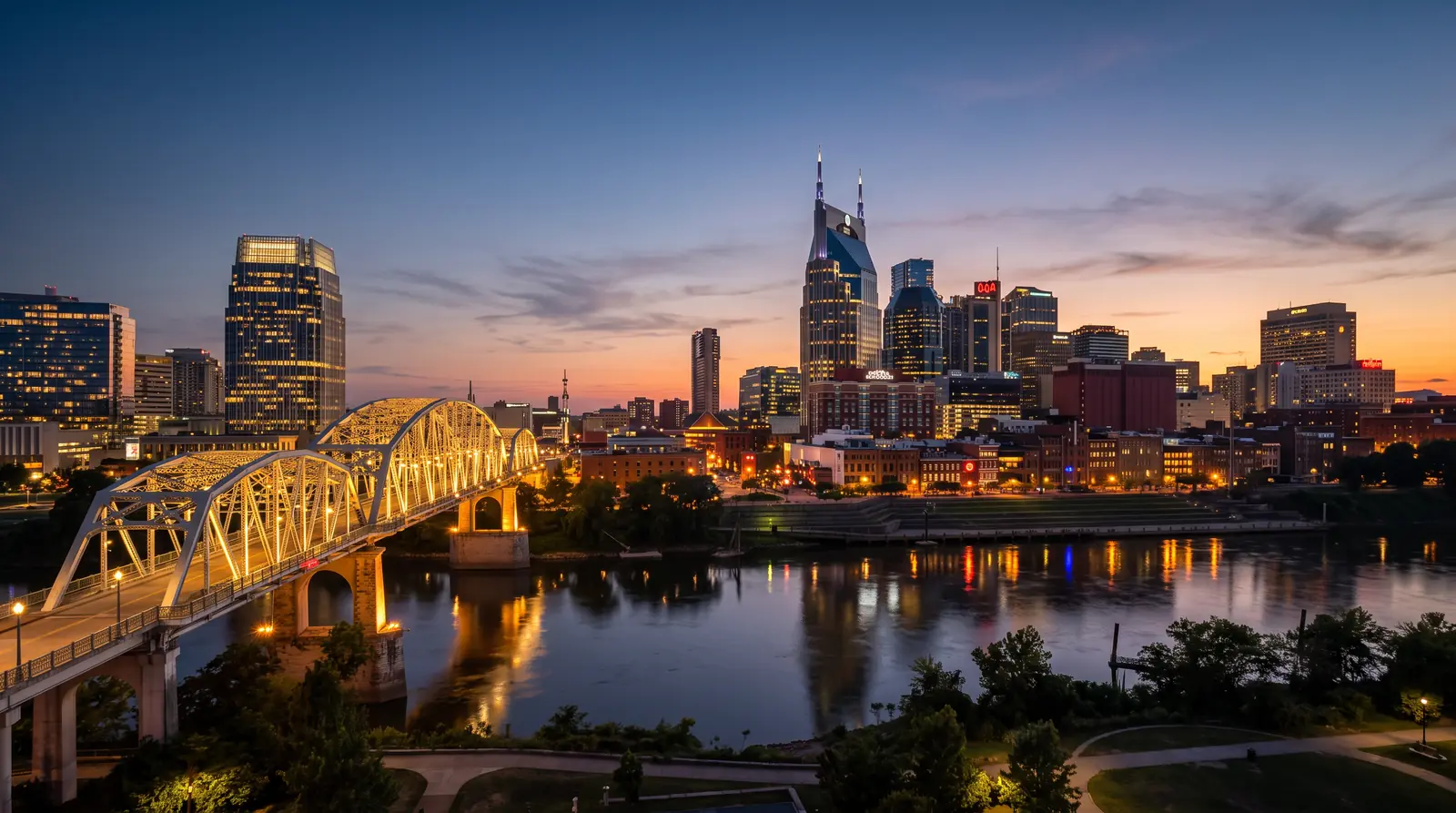 The Nashville skyline glowing at blue-hour dusk beyond the Cumberland River with a calm reflection, a warm sunset gradient in the sky and the lit pedestrian bridge in the foreground