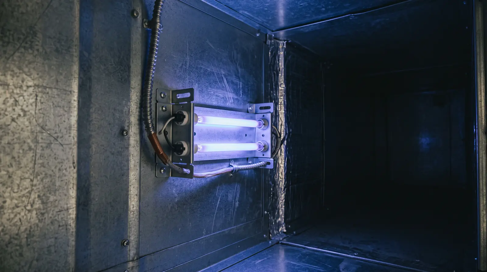 A compact ultraviolet lamp assembly mounted inside the plenum of an air handler glowing a soft violet-blue, clean metal surfaces, dark surroundings