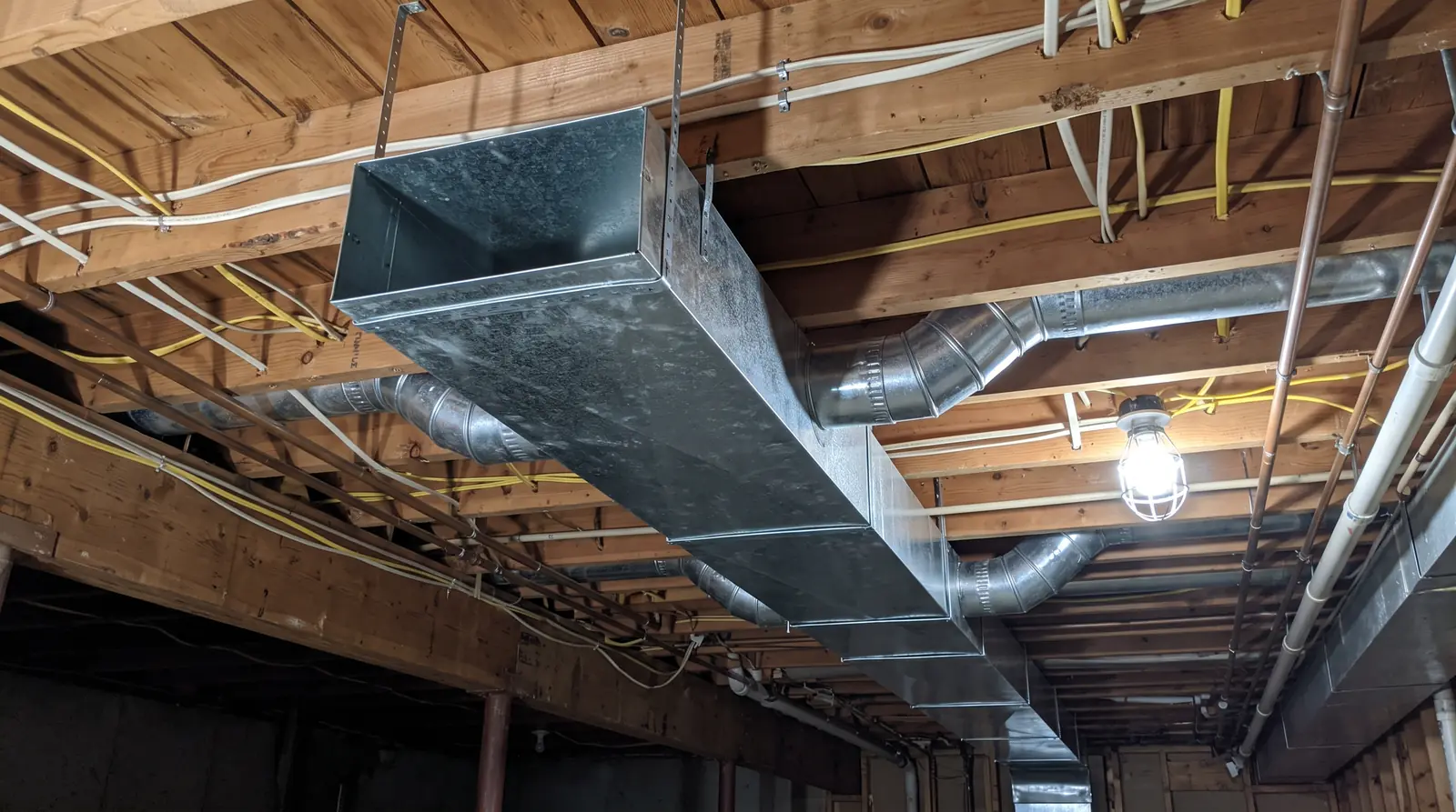 A galvanized sheet-metal main trunk duct with round branch takeoffs hung neatly from open basement joists with metal straps, bright LED work lights